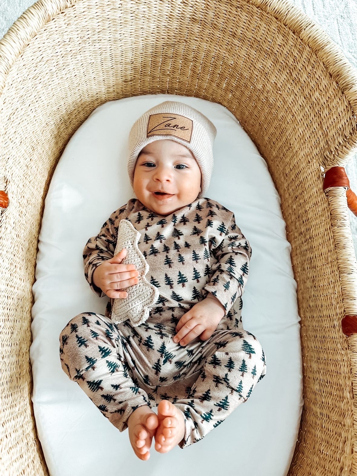 A baby dressed in a beige outfit with green pine tree prints and a light beige knit hat lies in a wicker bassinet. The baby is smiling while holding the Beige | Knit Tree Rattle from Forever French Baby and has their feet playfully touching. The Forever French Baby bassinet rests on a light-colored surface.