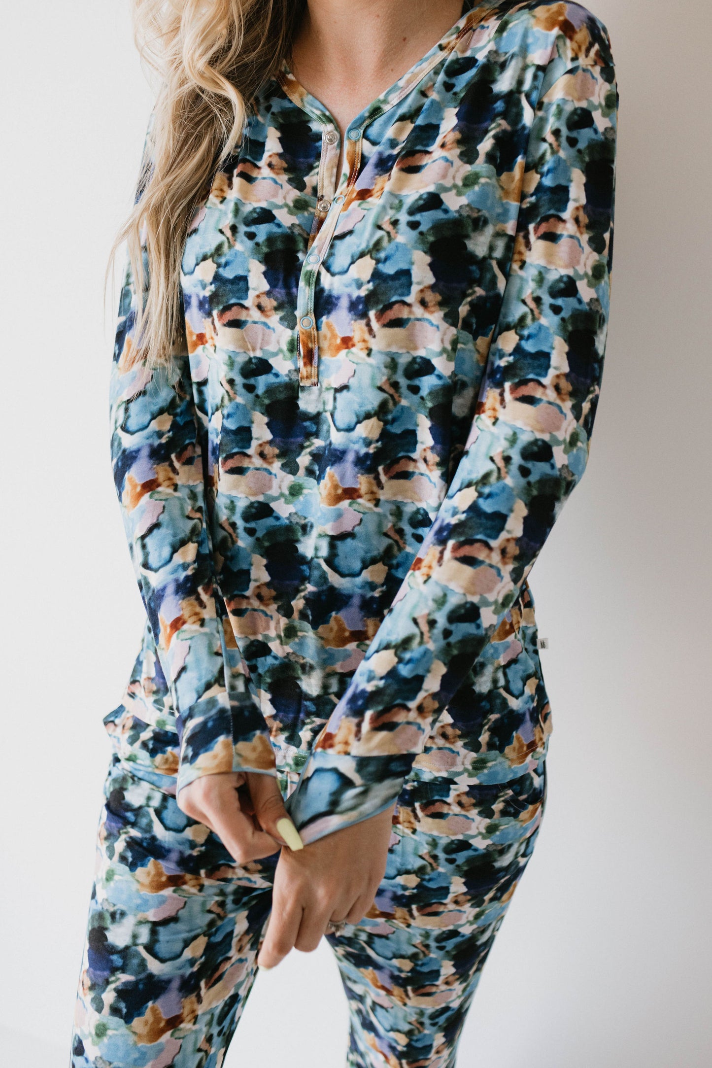 A person is standing against a plain white background, wearing the Women's Bamboo Pajamas in the Charli Print from forever french baby. The matching long-sleeve top and pants, featuring an abstract multicolored pattern with shades of blue, green, orange, and white reminiscent of Charli French designs, obscure their face in the image.