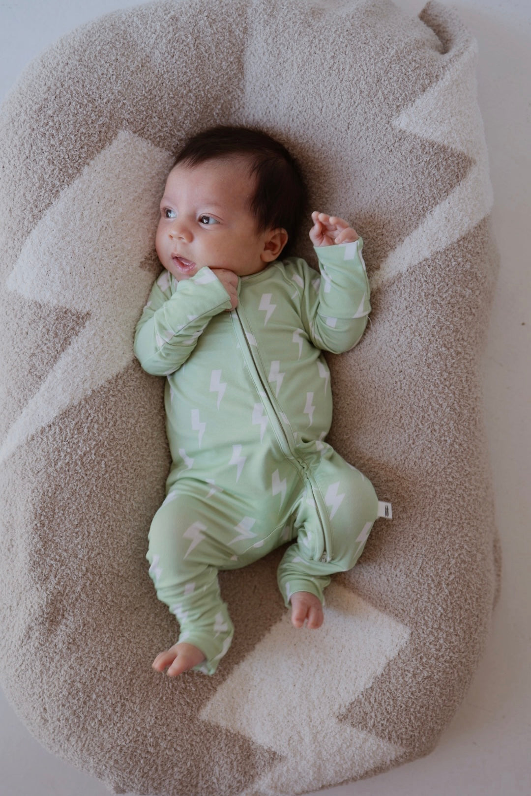 A baby in forever french baby's Bamboo Zip Pajamas | Flash—a mint green onesie with white lightning bolts—lies on a soft, beige cushion with white geometric patterns, gazing to the side.