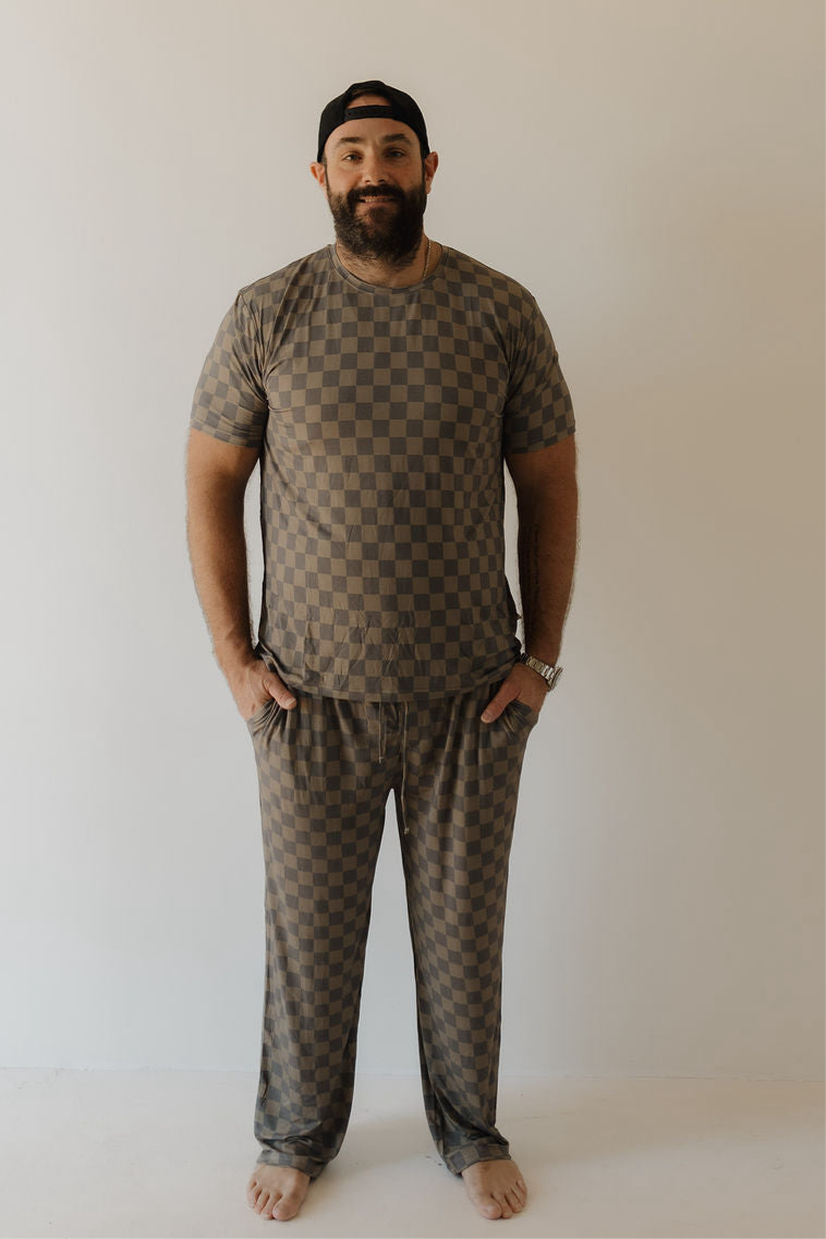 A bearded man in a backward black cap smiles with hands in his pockets, barefoot against a light backdrop, showcasing the comfort of forever french baby's Men's Bamboo Short Sleeve Pajama in Faded Checkerboard—soft, hypo-allergenic sleepwear.