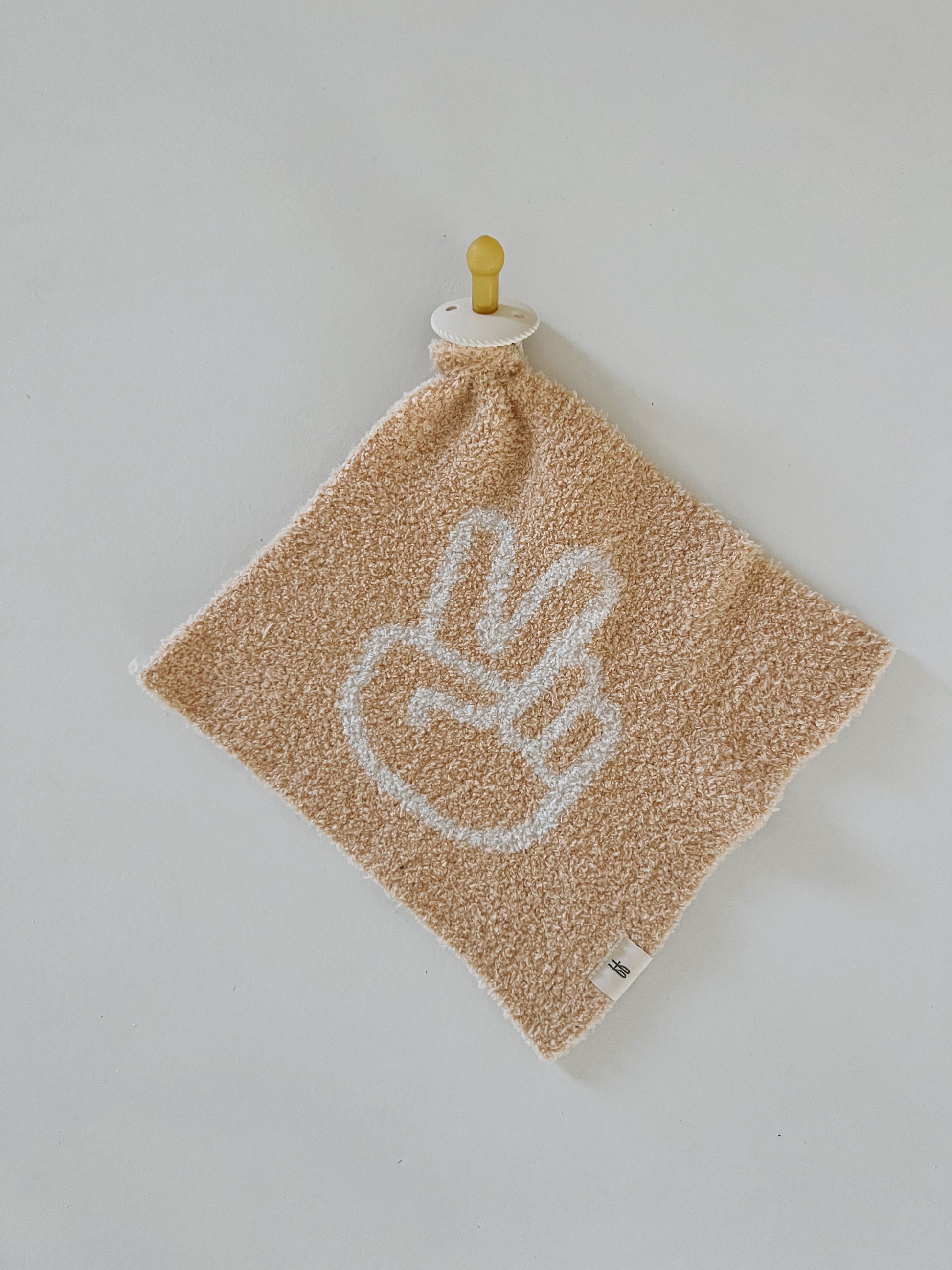 A Lovey | Peace Sign towel from forever french baby, featuring a beige, textured design with a white peace sign hand gesture in its center, hangs on a yellow knob against a plain white wall, evoking the spirit of a peaceful warrior.