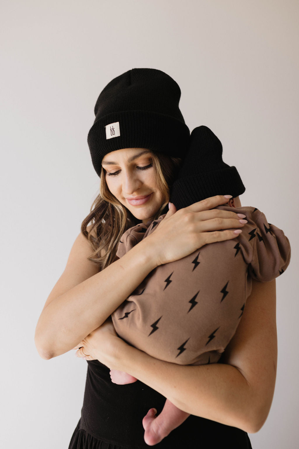 A woman in a black beanie gently hugs a baby dressed in the forever french baby Long Sleeve Romper | Lightning Bolt with a matching beanie. Both have their eyes closed, sharing an affectionate moment.