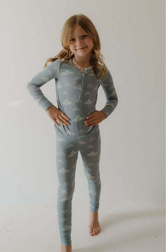 A young child with long blonde hair stands barefoot on a white floor, smiling at the camera with hands on hips, wearing Kendy 3’s Bamboo Two Piece Pajamas | Duke—light blue pajamas with a cloud pattern made from breathable fabric for all-night comfort.