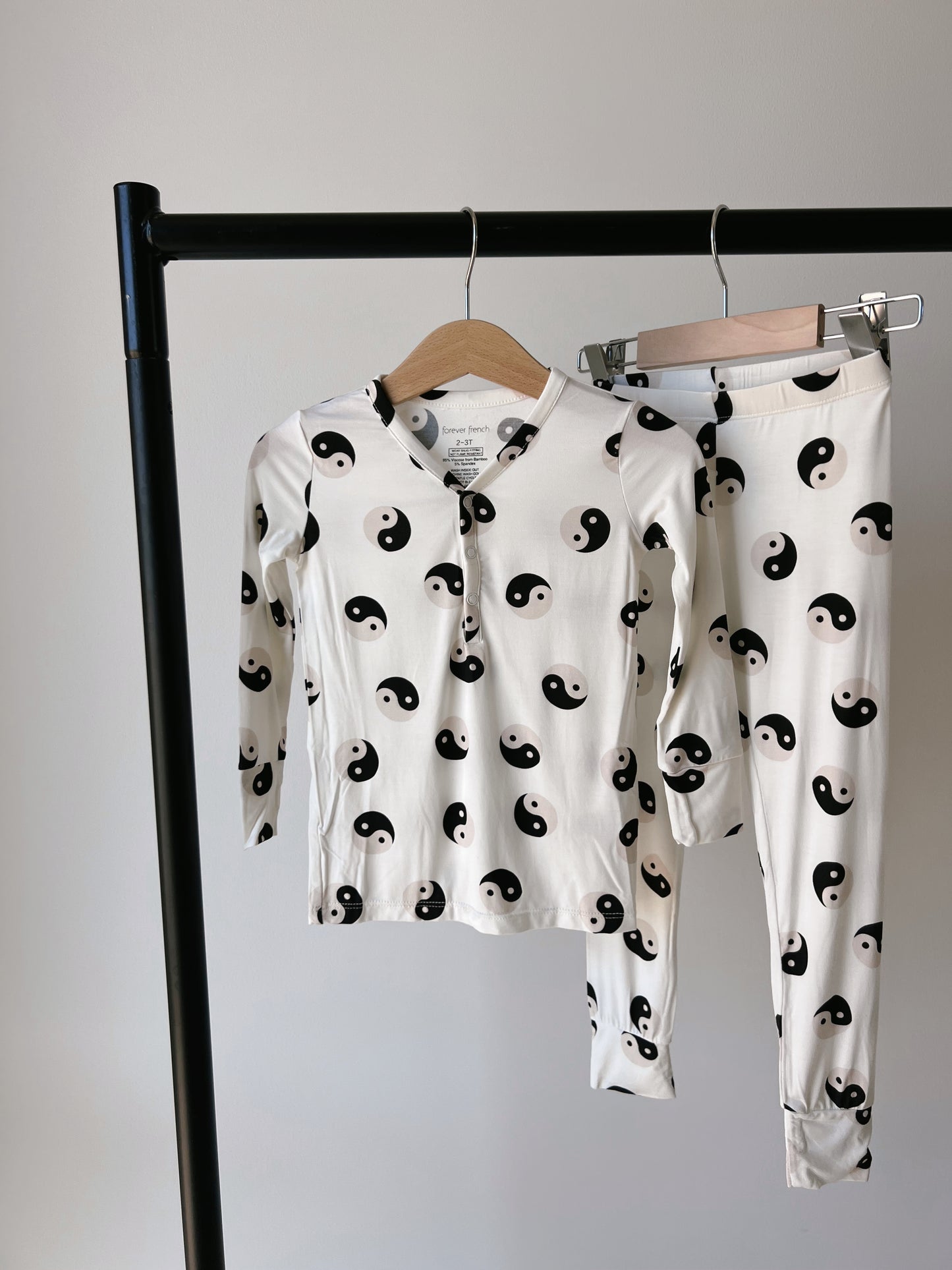 The Bamboo Two Piece Pajamas | Black & White Yin Yang by forever French baby are made from hypo-allergenic bamboo fabric. The set features a white design with black yin-yang symbols and includes a long-sleeved button-up top and matching pants, both displayed on wooden hangers on a sleek black clothing rack against a plain white background.
