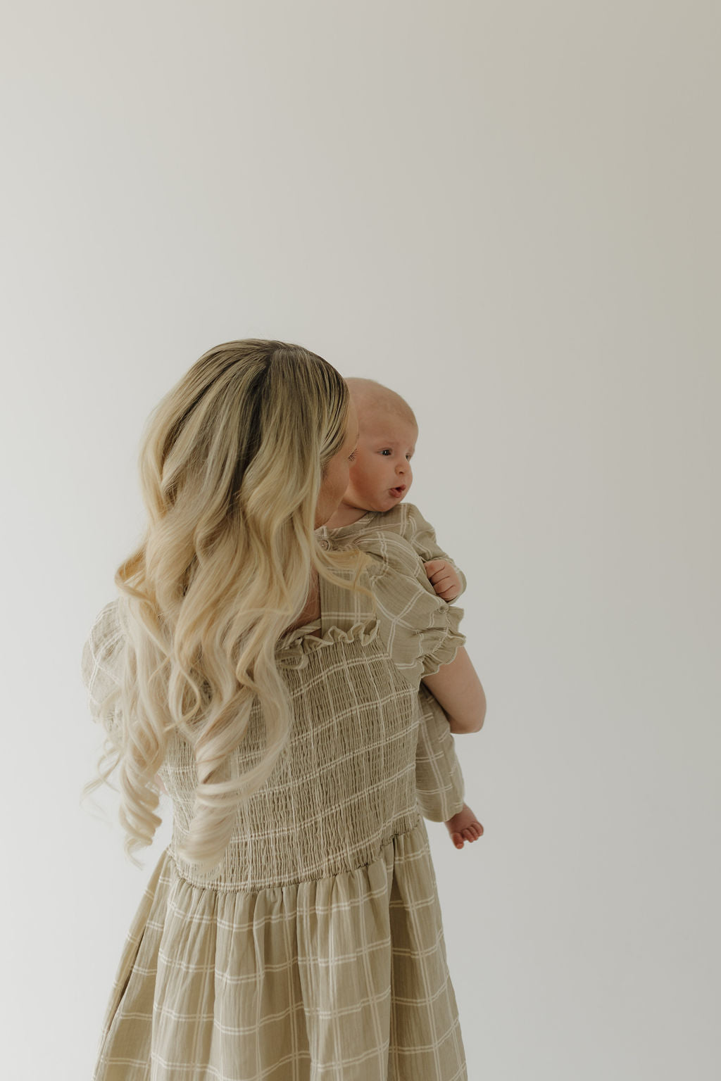 A woman with long, wavy blonde hair holds a baby against her shoulder. Both wear the forever french baby Women's Short Sleeve Muslin Dress in Linen Grid, made from 100% cotton, facing away against a plain background.