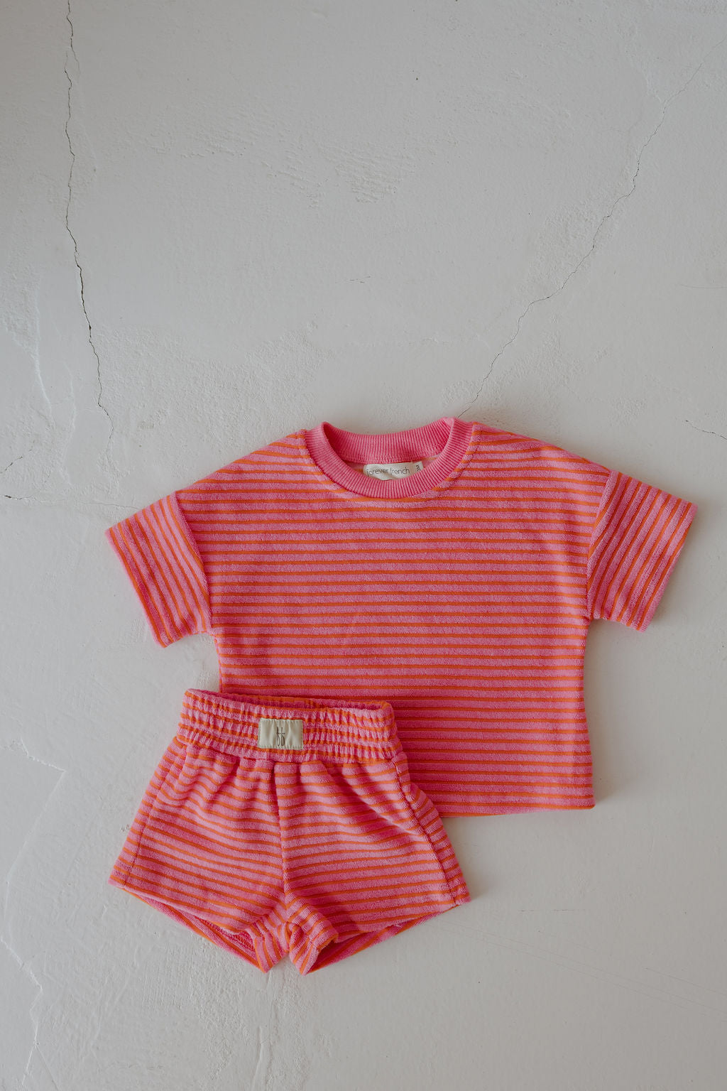 The forever french baby Child Terry Short Set | Strawberry features a matching short-sleeved shirt and shorts with pink and orange stripes, displayed flat on a light textured surface.
