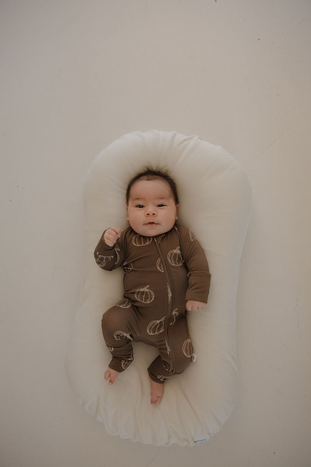 A baby in forever french baby's Bamboo Zip Pajamas | Brown Pumpkin lies on a soft, white, oval cushion against a plain light background.