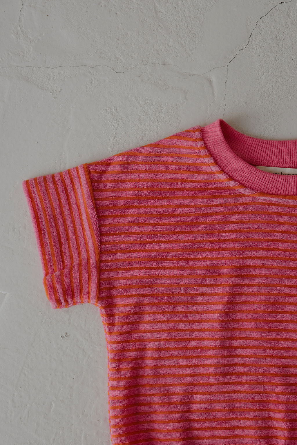 A close-up of the upper left of the forever french baby Terry T-Shirt Romper | Strawberry, featuring pink and orange stripes against an off-white background—designed for easy diaper changes and special family moments.