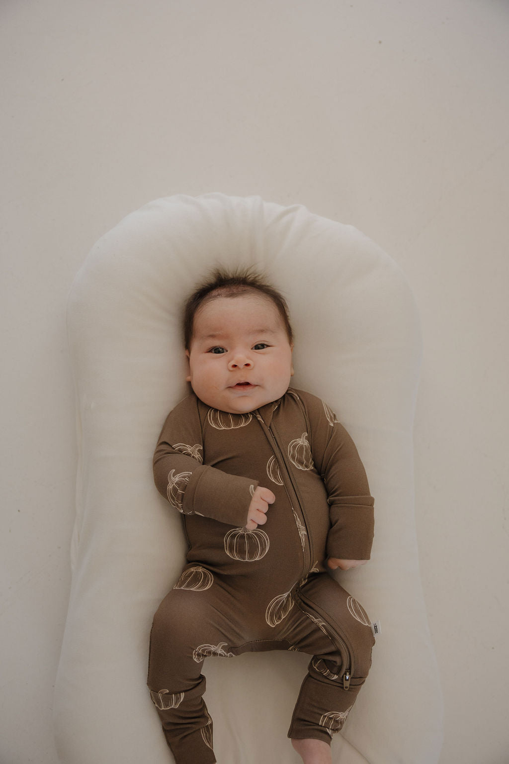 A baby lies on a white pillow, wearing forever french baby Bamboo Zip Pajamas in Brown Pumpkin, gazing upward calmly.
