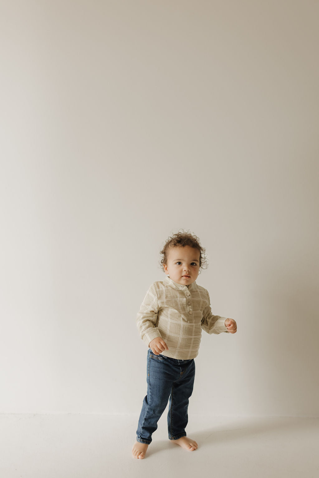 A young child with curly hair, wearing the forever french baby Muslin Button Up in Linen Grid, stands barefoot in blue jeans against a plain, light-colored background.