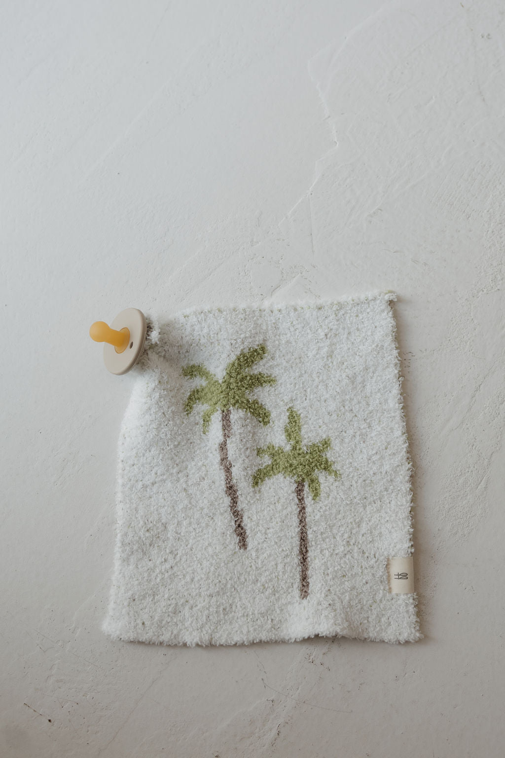 The Lovey | Palm by forever french baby features a yellow pacifier attached to a soft white cloth adorned with two green palm trees, shown lying on a textured white surface.