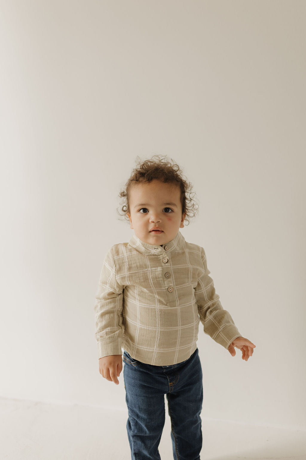 A young child with curly hair, wearing the forever french baby Muslin Button Up | Linen Grid shirt and blue jeans, stands against a plain light background, facing the camera.