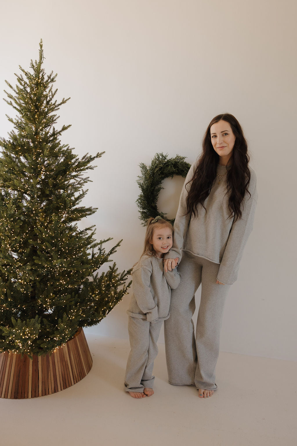 A woman and a young girl, both barefoot in the forever french baby Knit Pant Set | Grey (100% cotton), stand beside a Christmas tree with white lights, with a green wreath on the wall behind them.