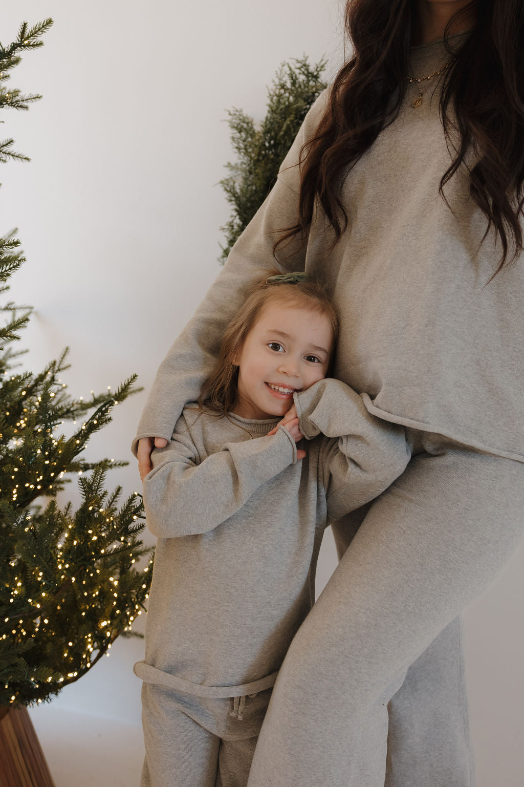 A smiling girl in a gray knit set and an adult wearing the Women's Knit Pant Set by forever french baby pose together by lit evergreen trees in a cozy indoor space, showcasing matching family outfits.