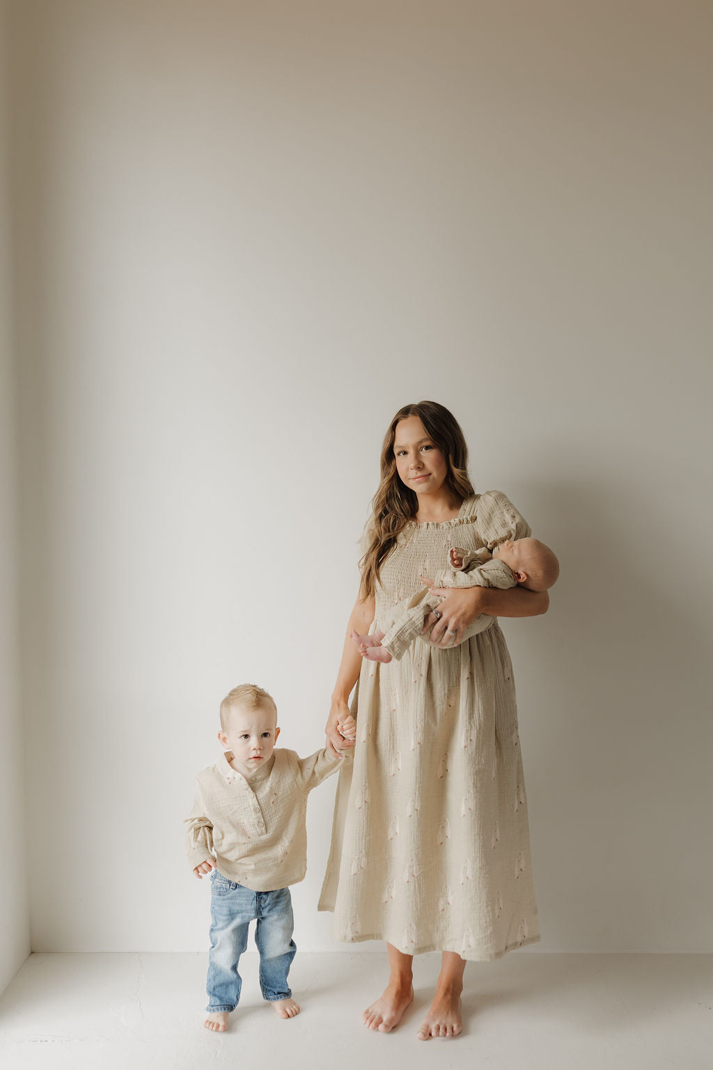 A woman in a beige Muslin Button Up by forever french baby stands barefoot in a minimal room, holding a baby and holding hands with a toddler dressed in a 100% cotton beige sweater and blue jeans.