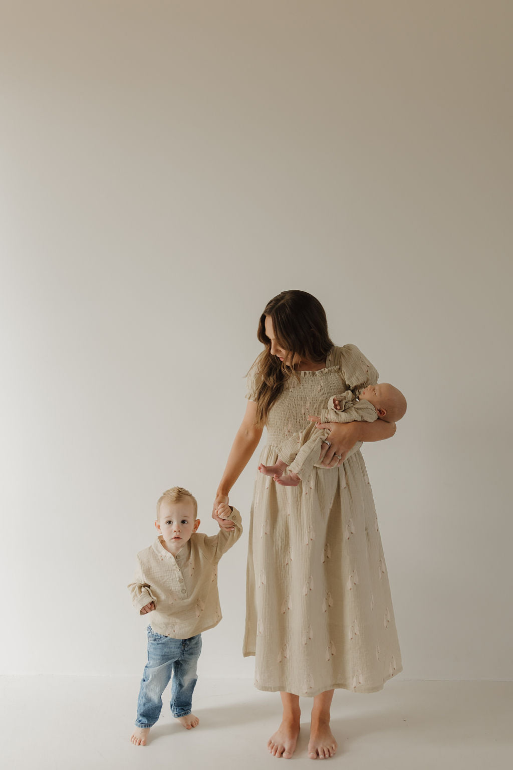 A barefoot woman in a beige dress holds a baby and stands beside a child wearing the forever french baby Muslin Button Up | Silly Goose, set against a plain, light-colored background.