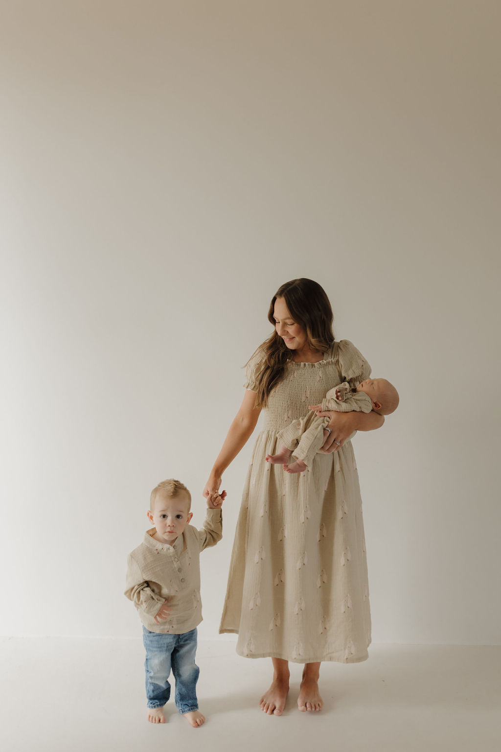 A woman in a beige dress stands barefoot, holding a baby and the hand of a young child dressed in the forever french baby Muslin Button Up | Silly Goose, all set against a plain light background.
