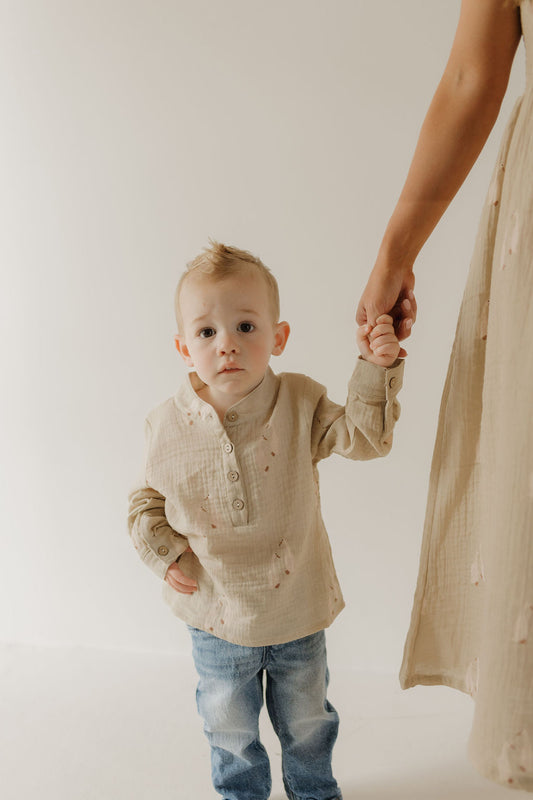A young boy with short blond hair wears the forever french baby Muslin Button Up in beige, holding hands with an adult in similar muslin styles. Both stand against a plain light background.