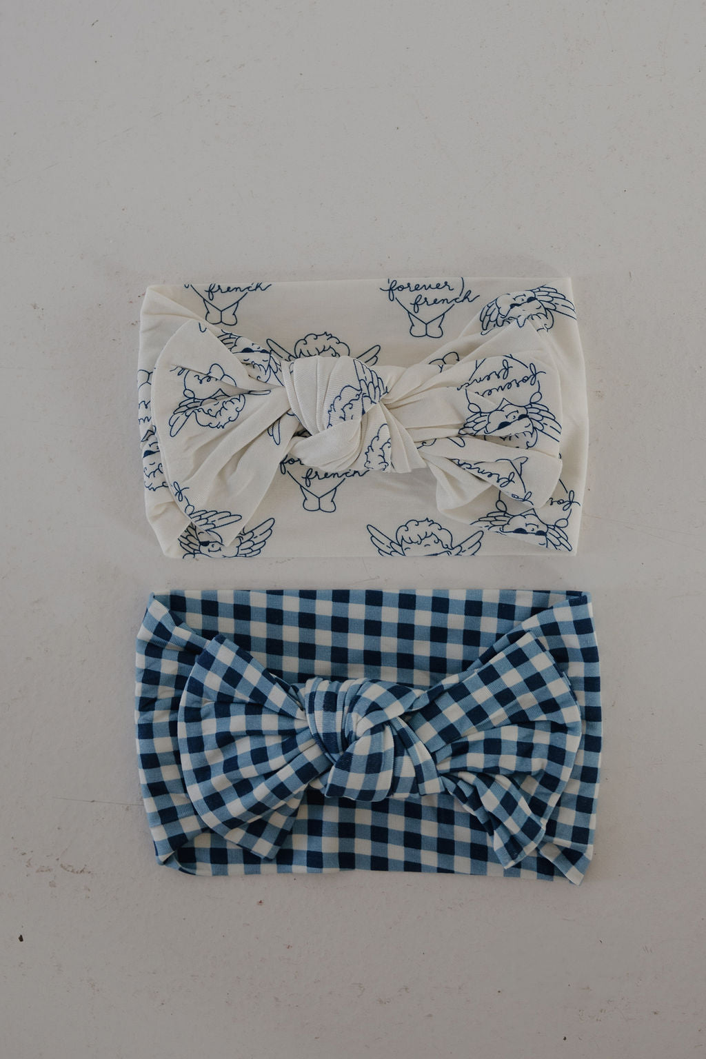 Two forever french baby bamboo head wraps: one in white with blue cherub sketches and script, the other in blue gingham. Both adjustable bow styles from the Bamboo Head Wrap | Blue Gingham collection.