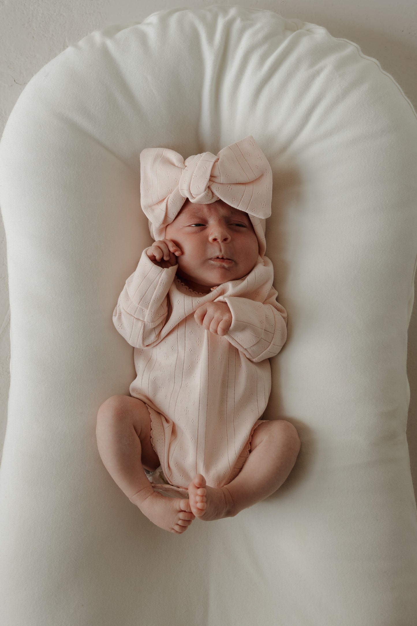 A baby wearing the forever french baby Long Sleeve Snapsuit | Ballerina in pale pink pointelle cotton, paired with a large matching bow headband, lies on a white cushioned surface, looking up with one hand near their face and legs slightly bent.