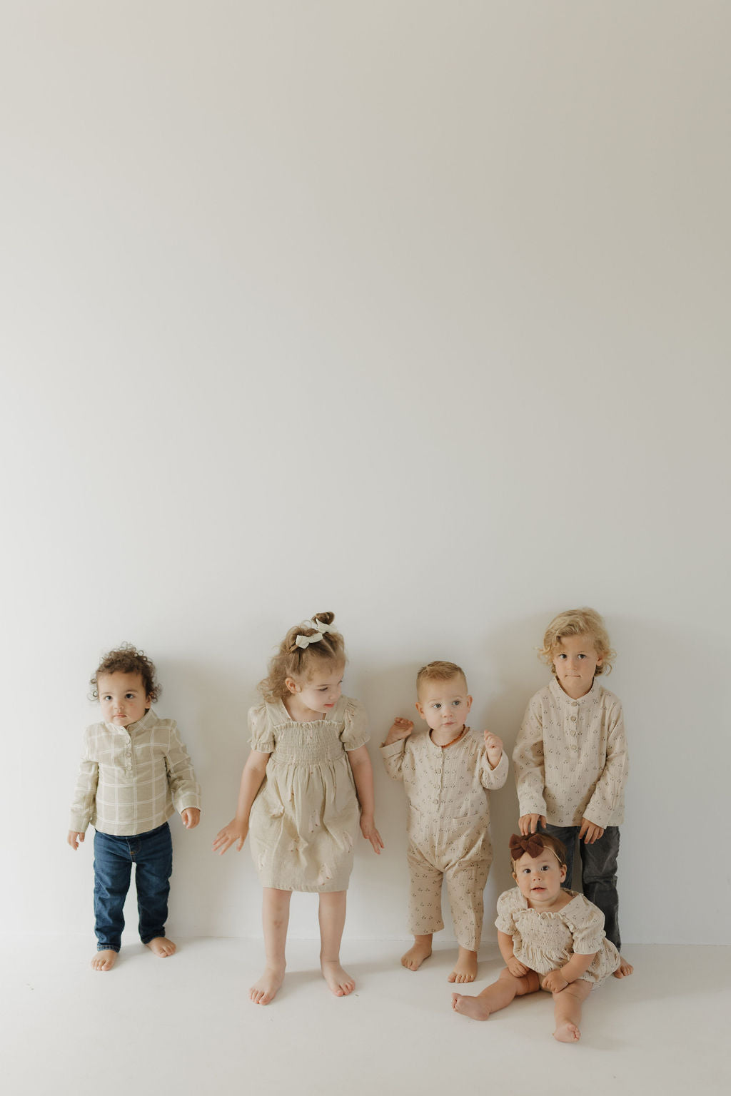 Four children stand and one sits against a white wall wearing neutral, 100% cotton outfits. The mood is calm and minimalistic. Featured: the Toddler Muslin Dress by forever french baby from Silly Goose.