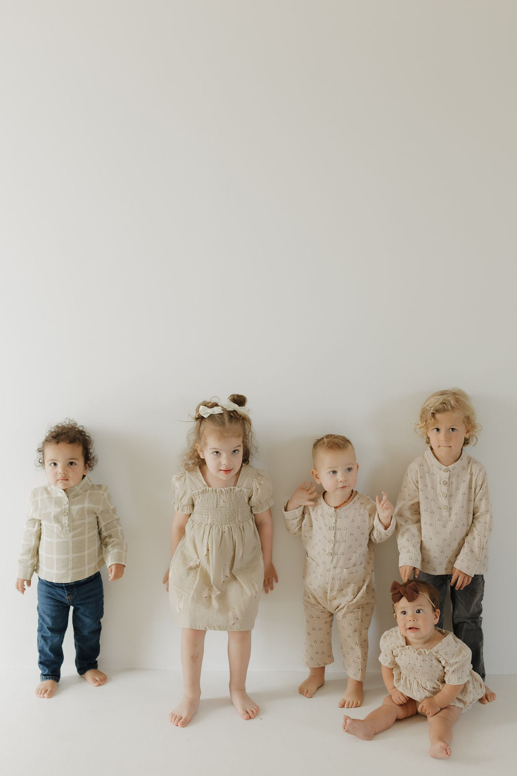 Five young children wear forever french baby's Toddler Muslin Dress | Silly Goose in neutral tones. Four stand and one sits before a bright white wall, the minimal background highlighting their 100% cotton outfits and joyful expressions.