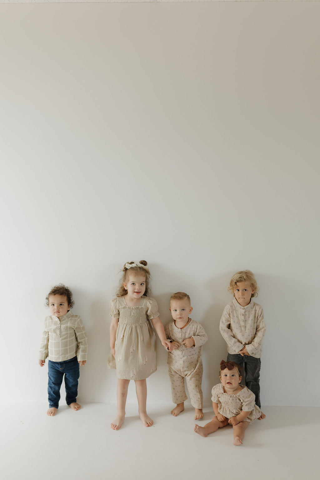 Five young children in neutral outfits, including the forever french baby Toddler Muslin Dress | Silly Goose made of 100% cotton, pose against a plain light wall. Four stand while one sits, creating a calm atmosphere with breathable fabrics.