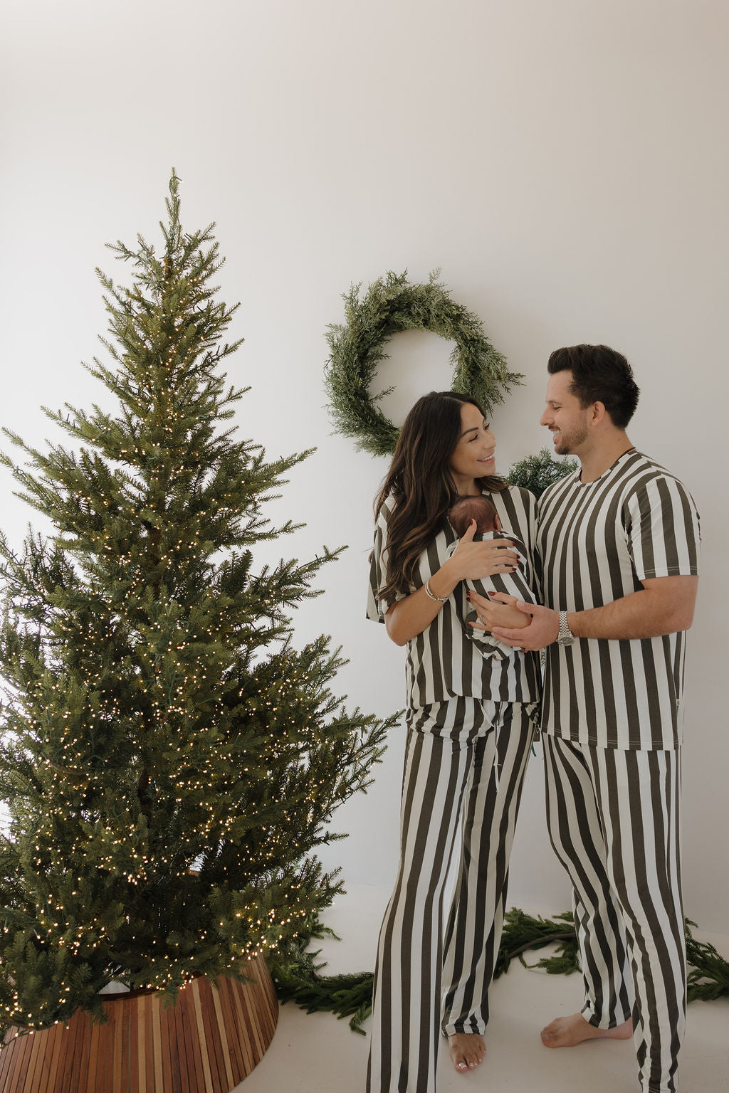 A smiling couple poses in front of a Christmas tree and green wreath, wearing forever french baby's Men's Short Sleeve Bamboo Pajamas in Fireside Stripe, while holding their baby.