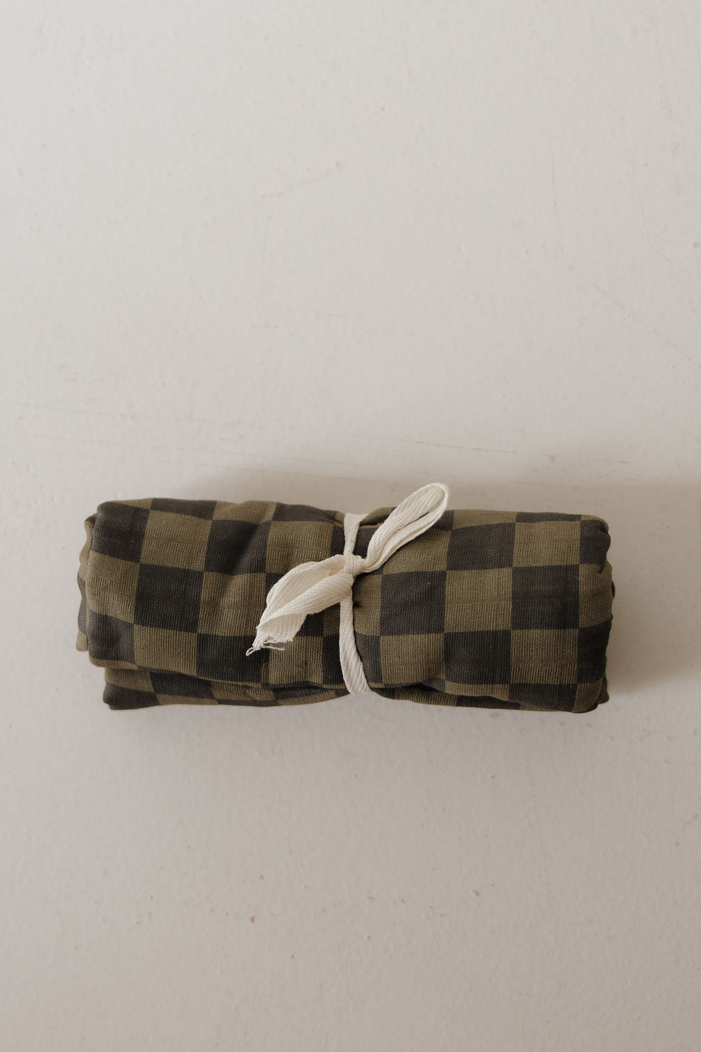 The Muslin Swaddle | Olive Checkerboard by forever french baby is a rolled-up muslin cotton swaddle with an olive checkered pattern, tied with white string and displayed on a light surface.