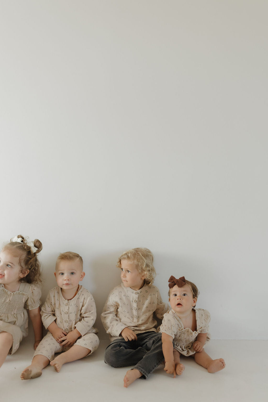 Four young children sit barefoot by a white wall in neutral outfits. One baby, wearing a brown bow, looks ahead in the forever french baby Toddler Muslin Dress | Silly Goose—made from 100% cotton muslin for all-day comfort and breathability.