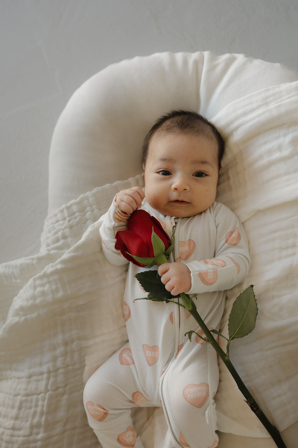A baby in forever french baby Bamboo Zip Pajamas | Love Notes - Grey lies on a soft white blanket, holding a red rose and looking at the camera, dressed in hypo-allergenic bamboo pajamas for ultimate comfort.