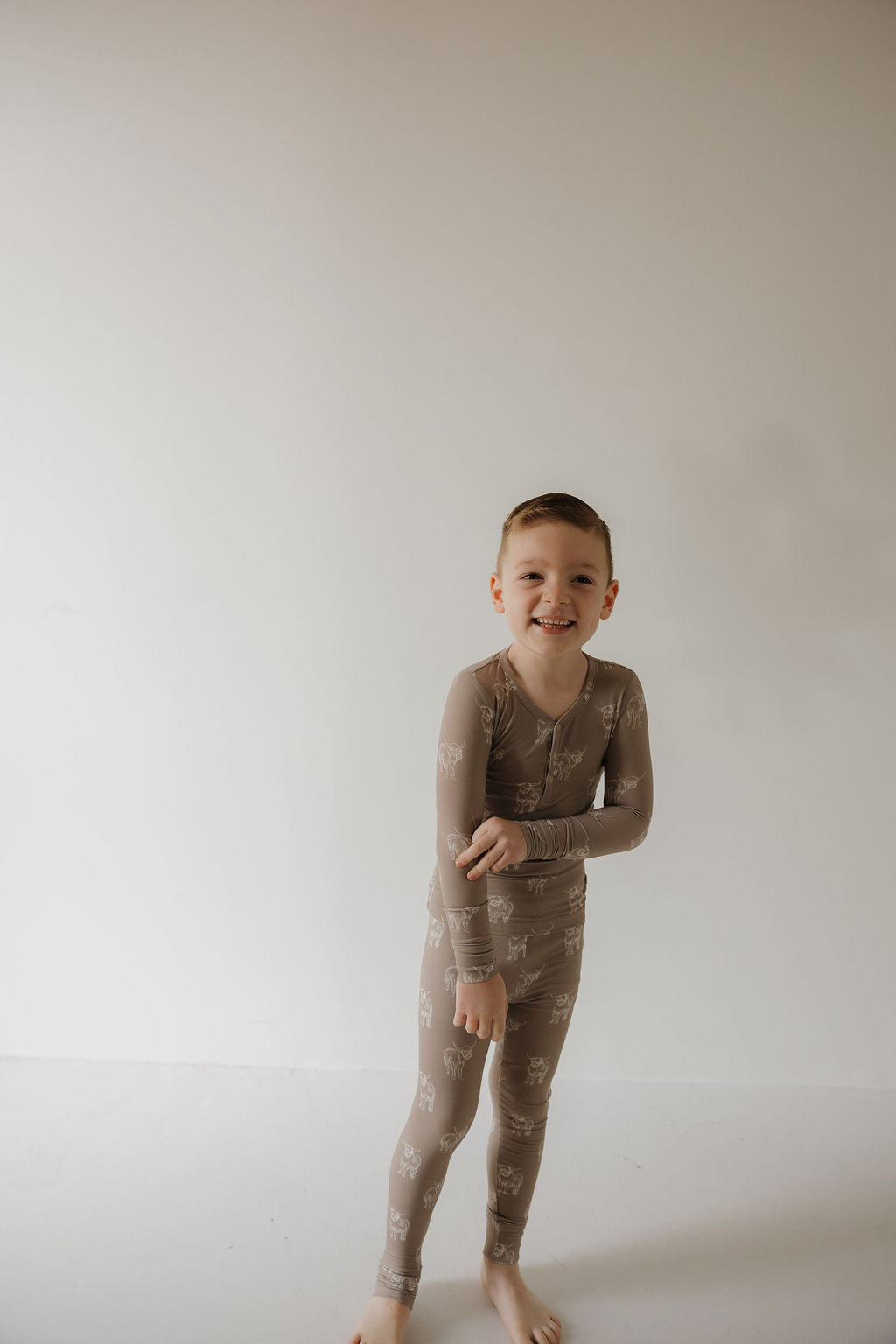 A young boy, barefoot and smiling, stands against a white wall wearing forever french baby's Bamboo Two Piece Pajamas | Highland—light brown, long-sleeved kids sleepwear with white animal prints, perfect for hypoallergenic comfort.