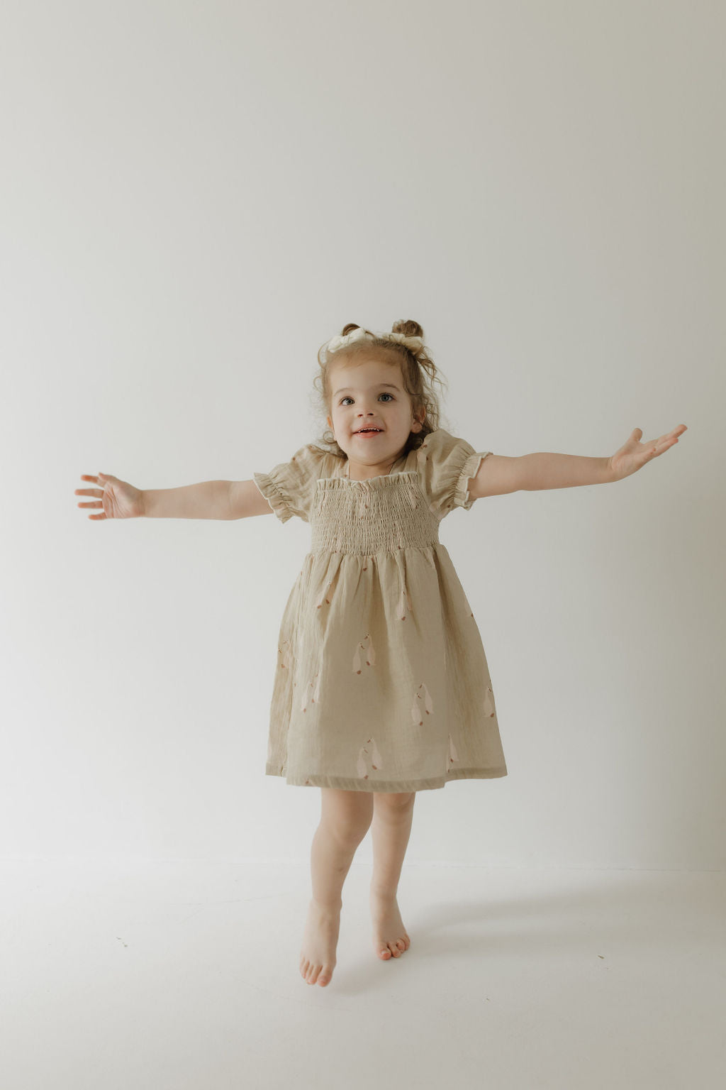A smiling girl poses barefoot with arms outstretched against a plain background, showcasing the breathable comfort of the Toddler Muslin Dress by forever french baby | Silly Goose.
