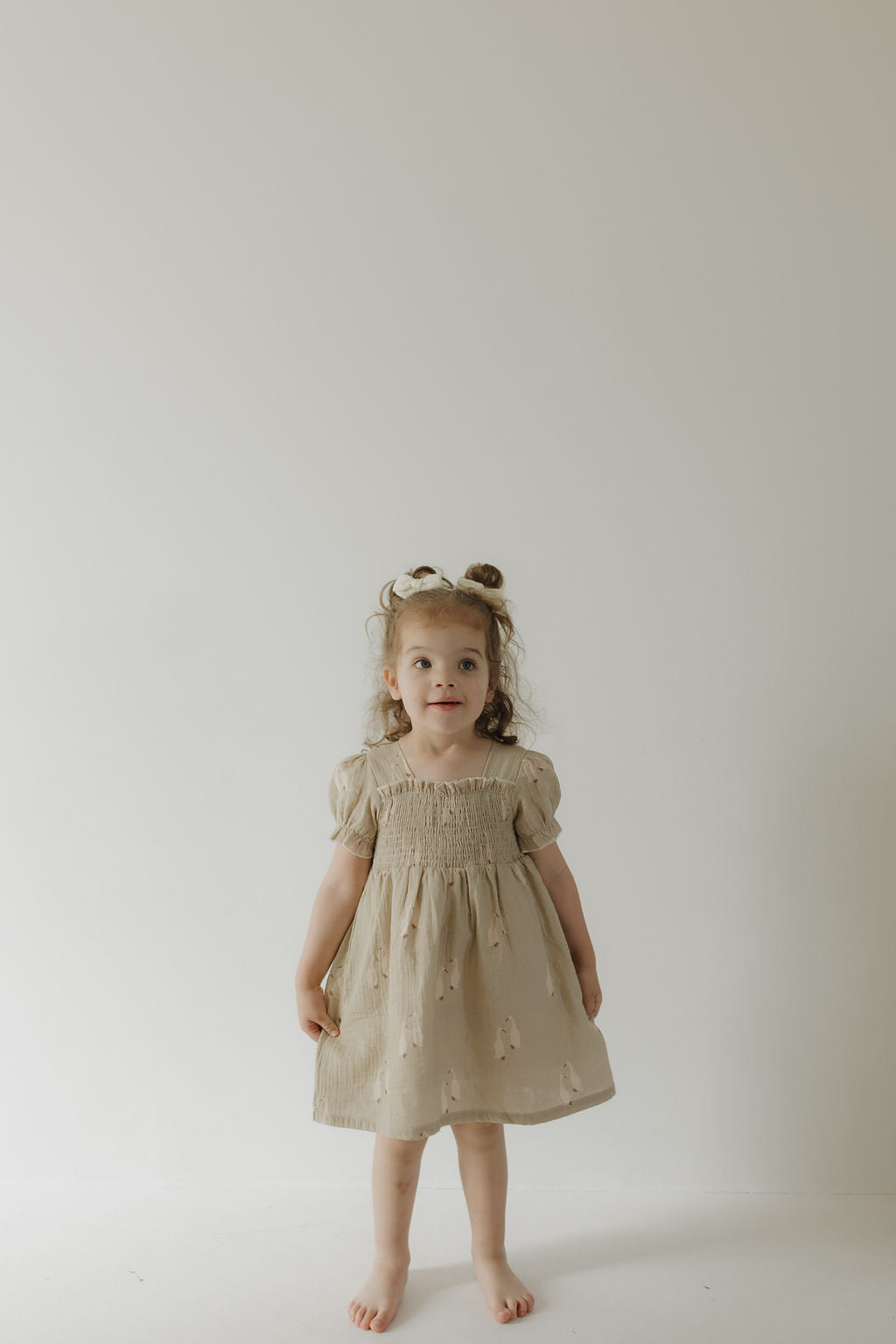 A young child with curly hair wears the forever french baby Toddler Muslin Dress | Silly Goose—a light beige, 100% cotton dress with puffed sleeves—standing barefoot against a plain, light background.