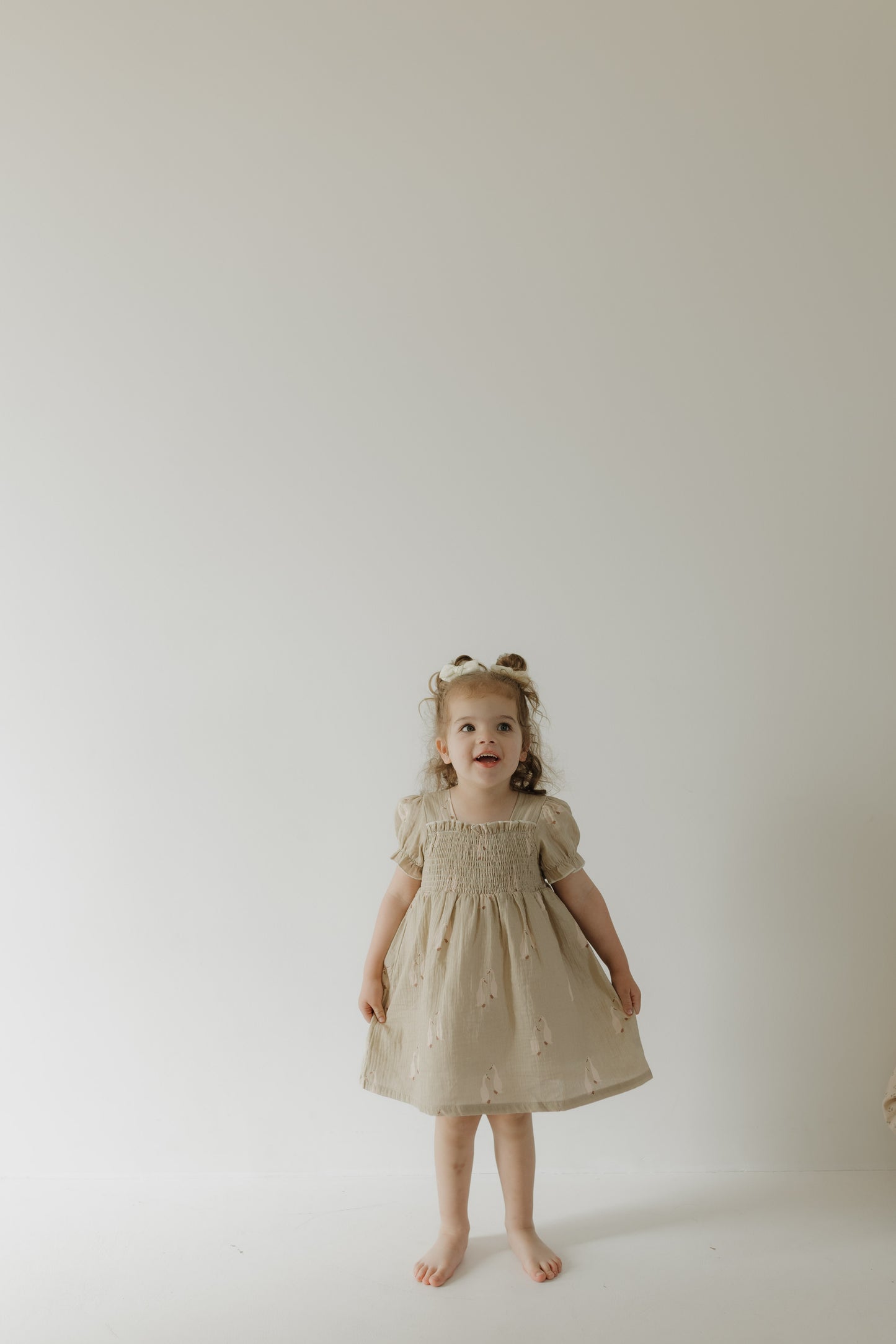A young child with curly hair stands barefoot against a light background, wearing the forever french baby Toddler Muslin Dress | Silly Goose with puffed sleeves, looking up with a cheerful expression.