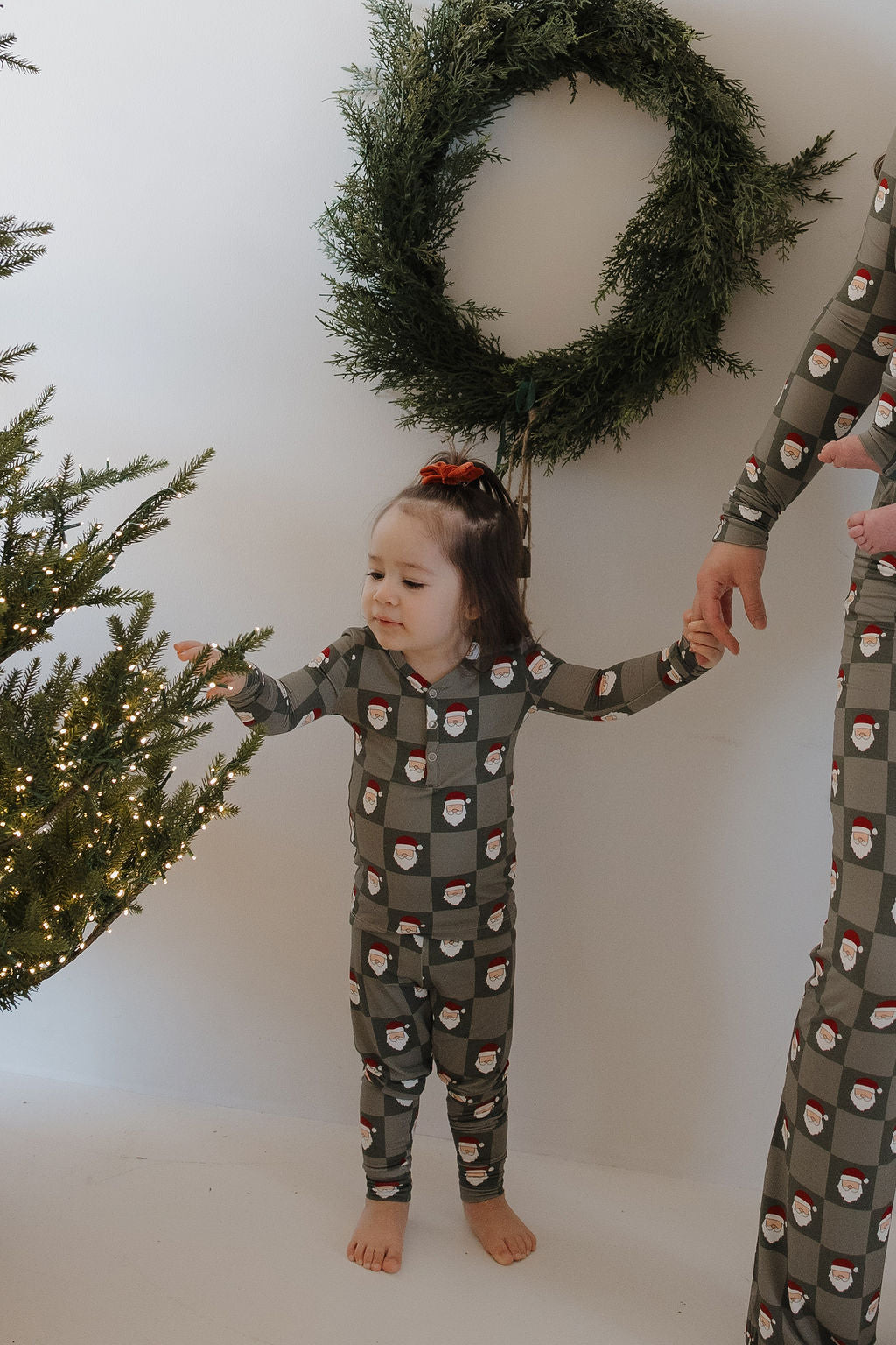 A young child in forever french baby Bamboo Two Piece Pajamas | Santa Checkerboard reaches toward a lit Christmas tree while holding an adult's hand. A green wreath decorates the white wall behind them.