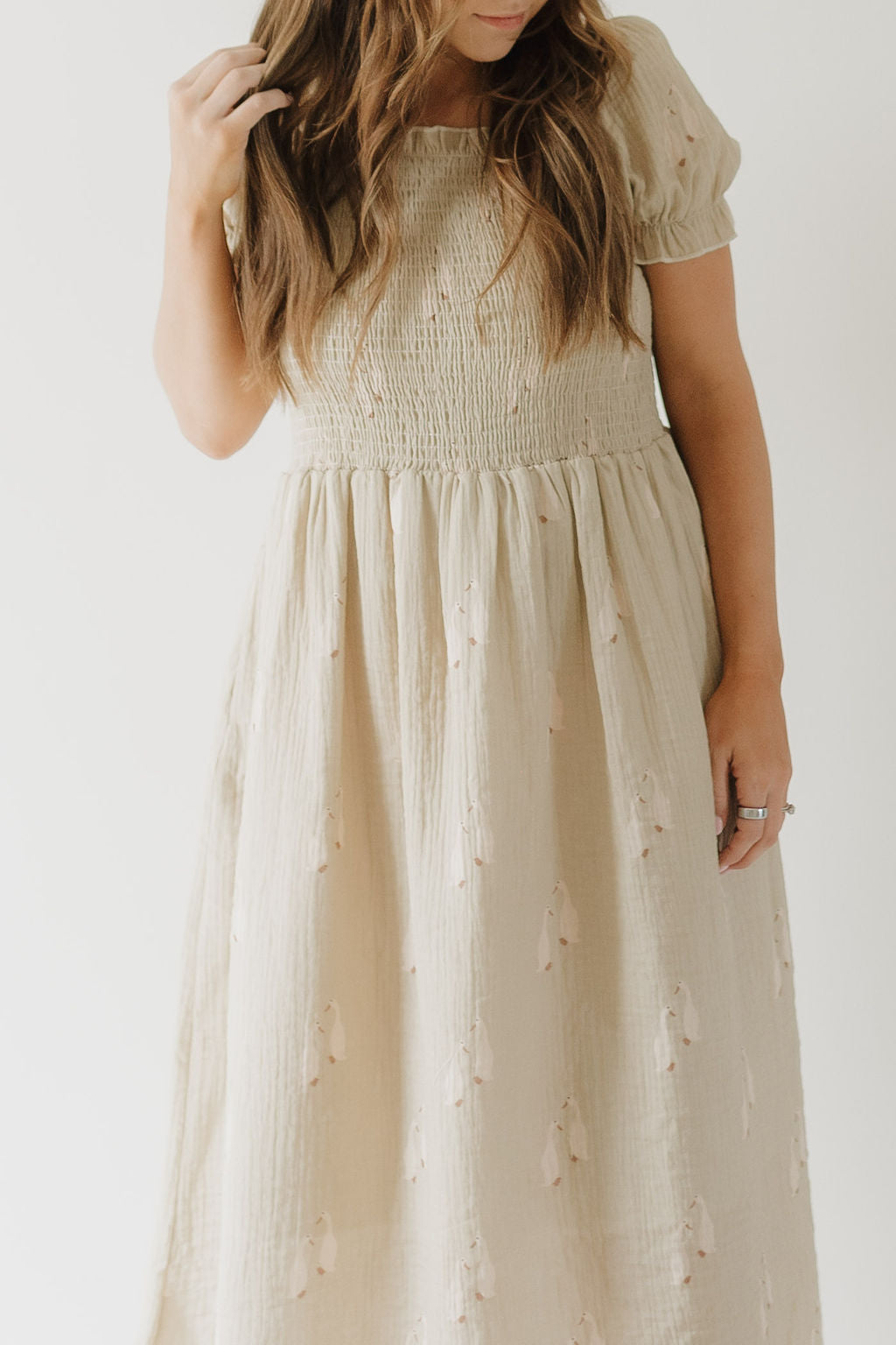 A woman with long brown hair models the forever french baby Women's Short Sleeve Muslin Dress | Silly Goose, a cream 100% cotton midi dress with short sleeves, smocked bodice, gathered waist, and subtle embroidery, standing against a plain background.