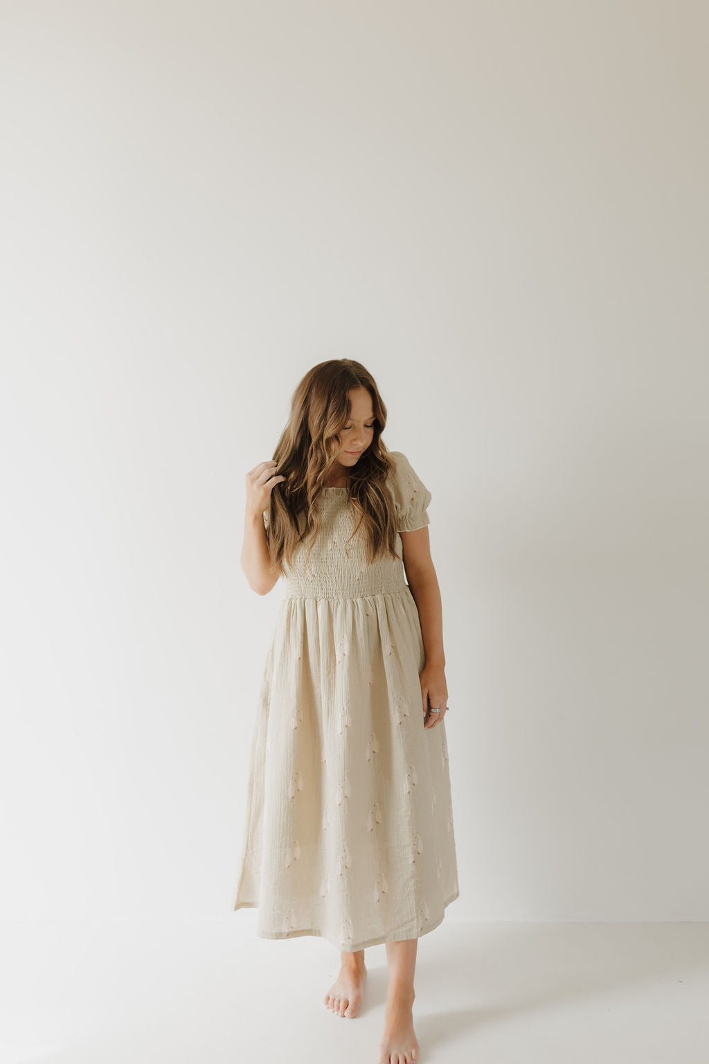 A barefoot woman with long hair stands against a plain light background, wearing the forever french baby Women's Short Sleeve Muslin Dress | Silly Goose. She looks down and touches her hair with one hand.