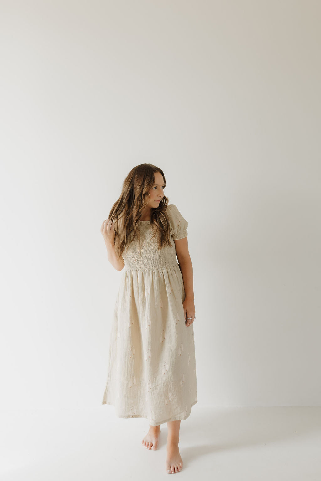 A woman with long brown hair stands barefoot against a white background, wearing the forever french baby Women's Short Sleeve Muslin Dress | Silly Goose in light beige, featuring a smocked bodice and midi skirt. Made from 100% cotton.