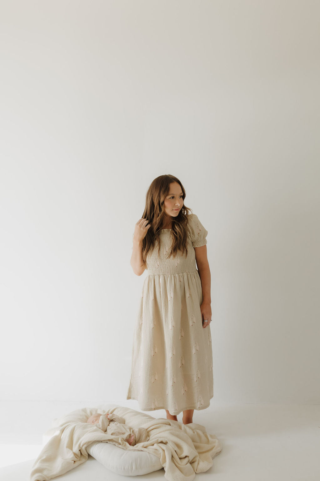 A woman wearing the forever french baby Women's Short Sleeve Muslin Dress by Silly Goose stands barefoot on a white floor, looking left. A baby in a blanket rests on a cushion at her feet, set against a simple light background.
