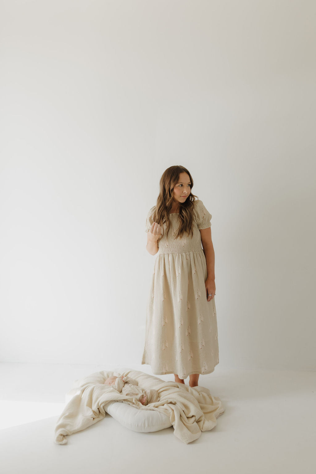A woman wears the forever french baby Women's Short Sleeve Muslin Dress by Silly Goose, barefoot on a white floor. A baby, swaddled in cream on a white pillow, lies before her against a plain, softly lit background.