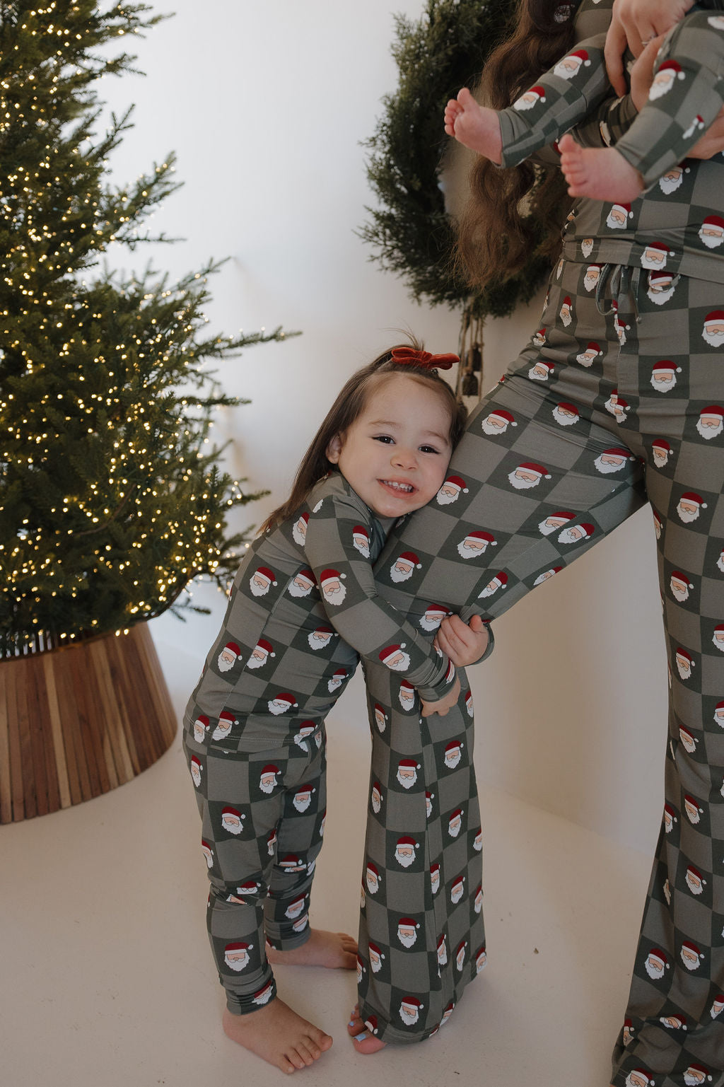 A young child in forever french baby Bamboo Two Piece Pajamas | Santa Checkerboard hugs an adult’s leg in matching sets, both standing by a Christmas tree and wreath in a cozy, festive scene.