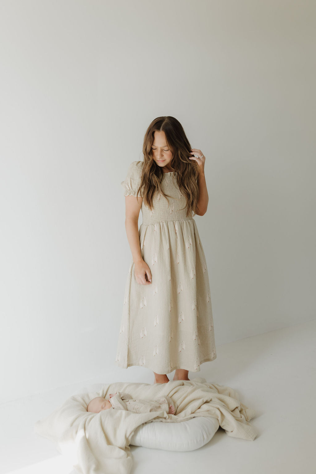 A woman wearing the forever french baby Women's Short Sleeve Muslin Dress by Silly Goose stands barefoot on a white floor, smiling as she looks down at a baby wrapped in a soft 100% cotton blanket on a cushioned blanket, against a plain light background.