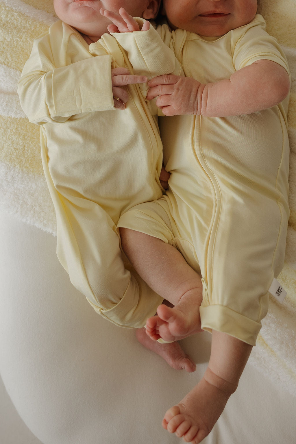 Two babies wearing matching “Sunny” Bamboo Zip Pajamas from forever french baby lie side by side on a soft, white blanket, their arms and legs gently overlapping. These hypoallergenic pajamas ensure they stay comfortable and cozy while they rest.