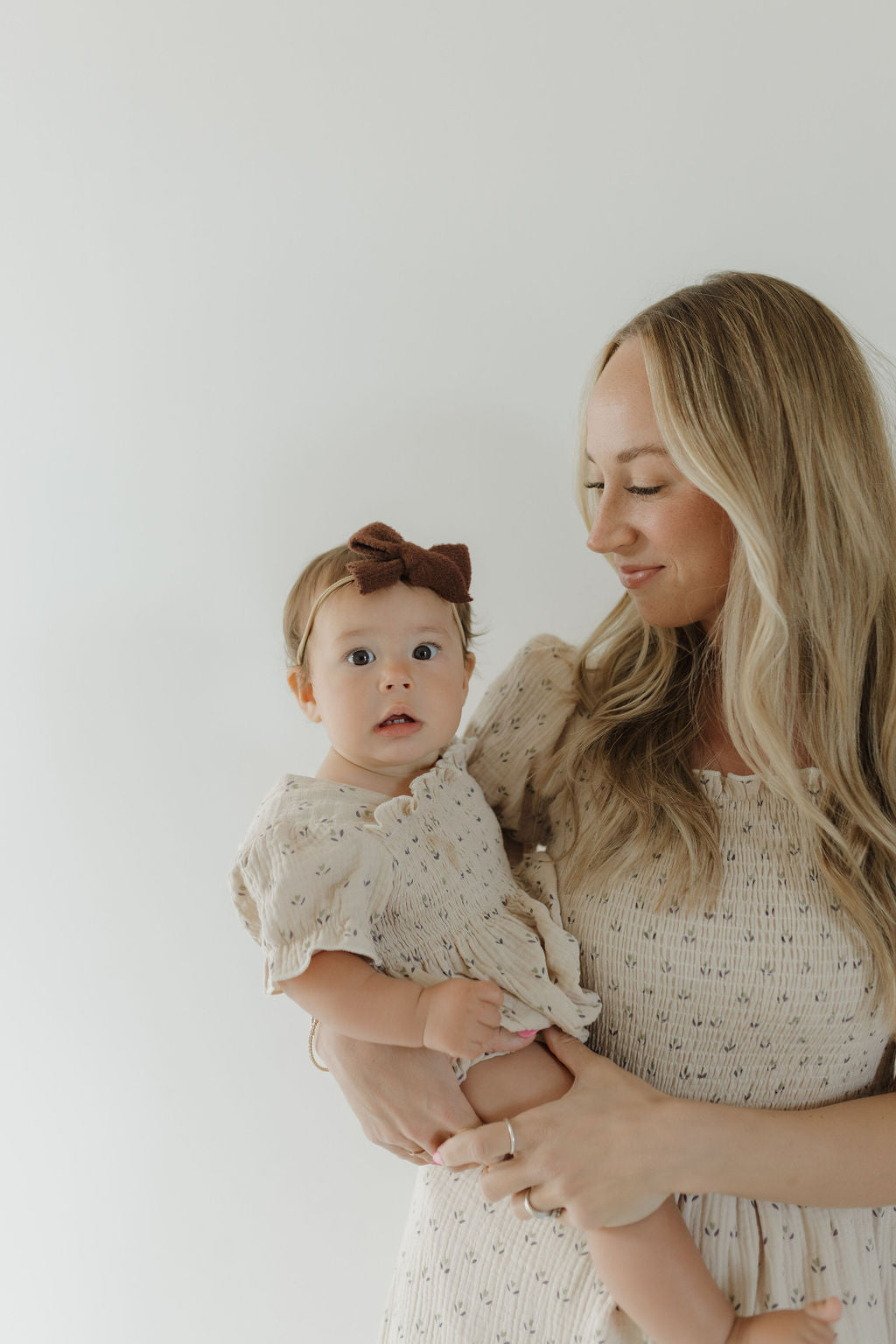 A woman with long blonde hair holds a baby in a brown bow headband. Both wear matching forever french baby Women's Short Sleeve Muslin Dress | Flora, made from lightweight cotton, standing against a plain white background.