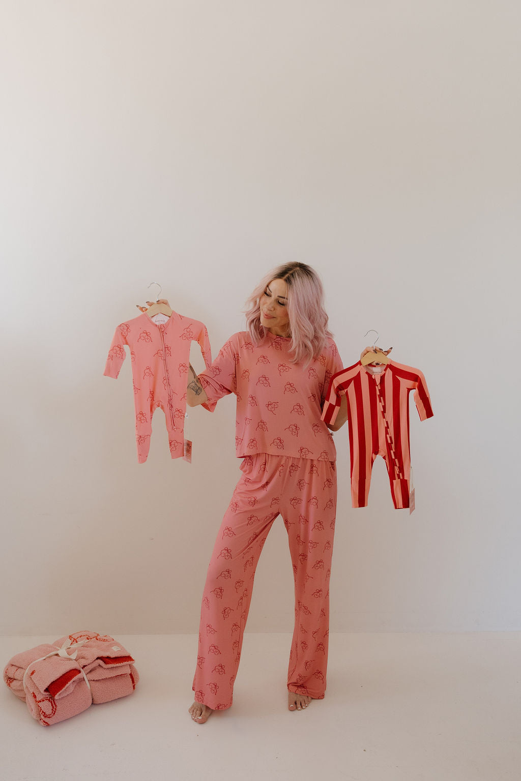 A woman with pink hair in pink pajamas stands barefoot indoors holding Minty Cherries Bamboo Zip Pajamas in Iconic Stripe and a pink variant, highlighting soft, hypo-allergenic baby clothing. Folded blankets rest by a plain white wall.
