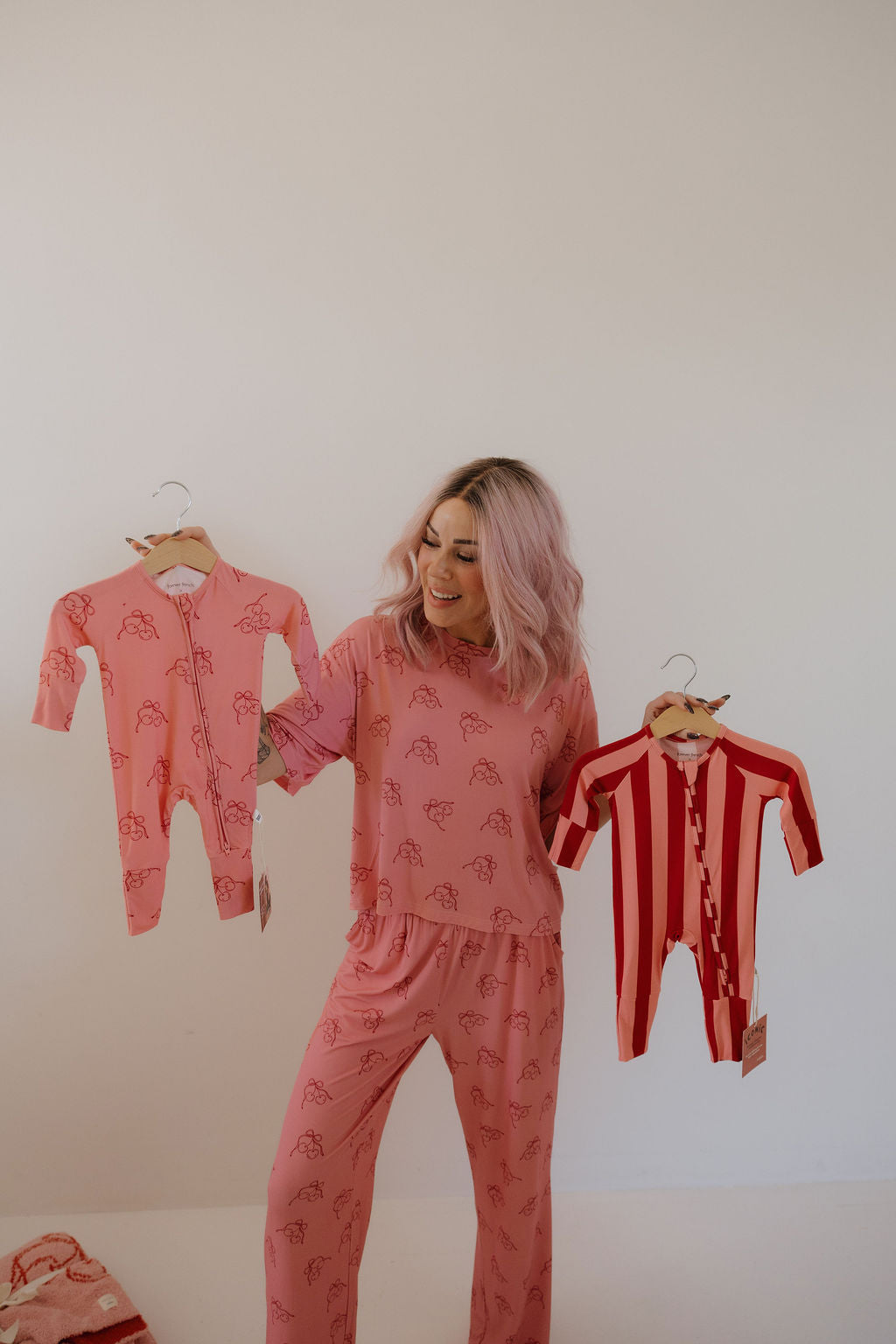 A woman with pink hair in Minty Cherries Bamboo Zip Pajamas | Iconic Cherry holds two baby onesies—one pink with turtles, one red and white striped—smiling at the breathable baby sleepwear against a plain white background.