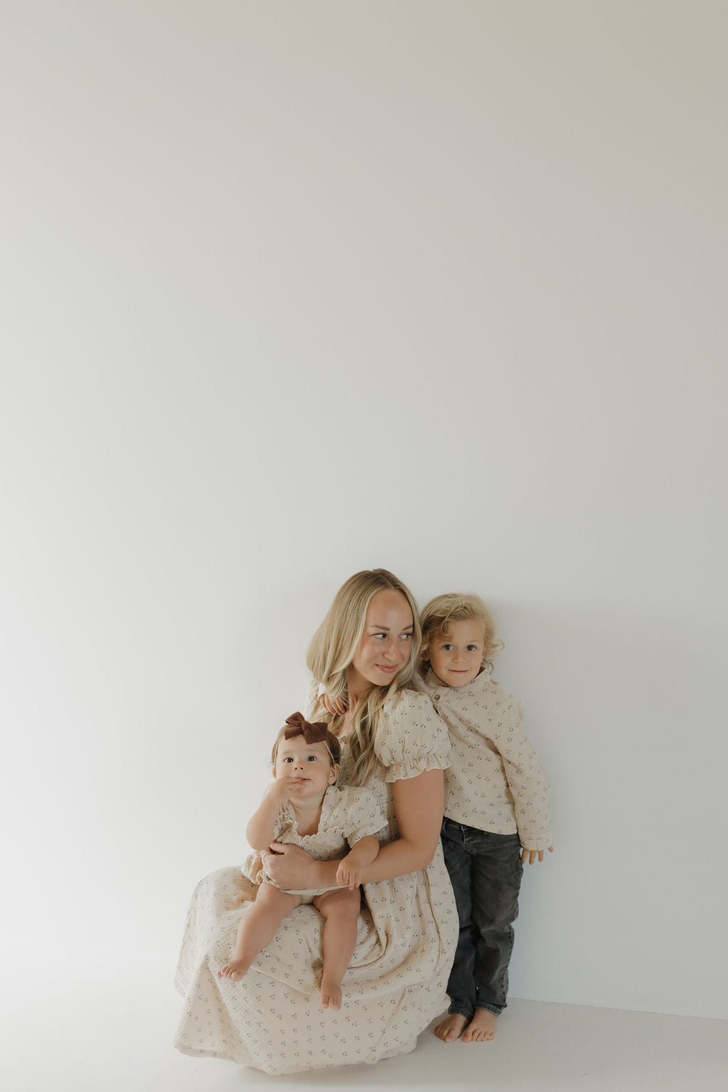 A woman wears the forever french baby Women's Short Sleeve Muslin Dress | Flora, sitting on the floor with a baby on her lap and a child beside her. All three are in coordinating light matching dresses against a plain white background.