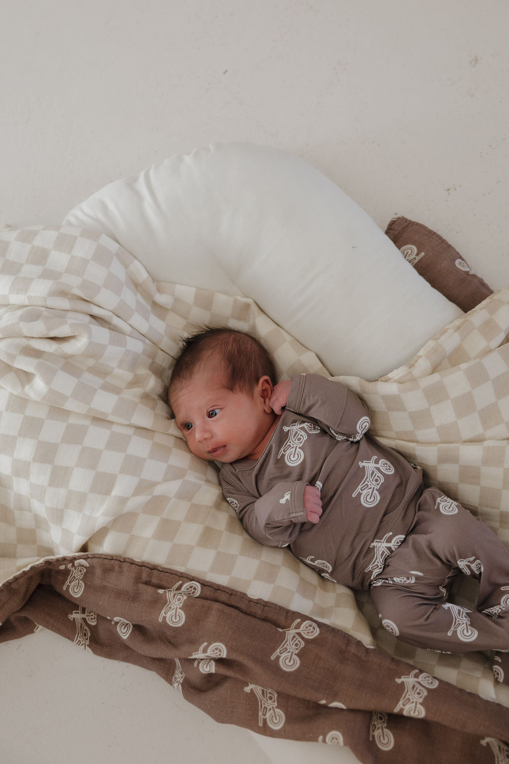 A newborn rests on a soft cushion, swaddled in a brown outfit with white details, beside the forever french baby Muslin Quilt | Dirt Bike & Checkerboard featuring cozy brown and beige checks.