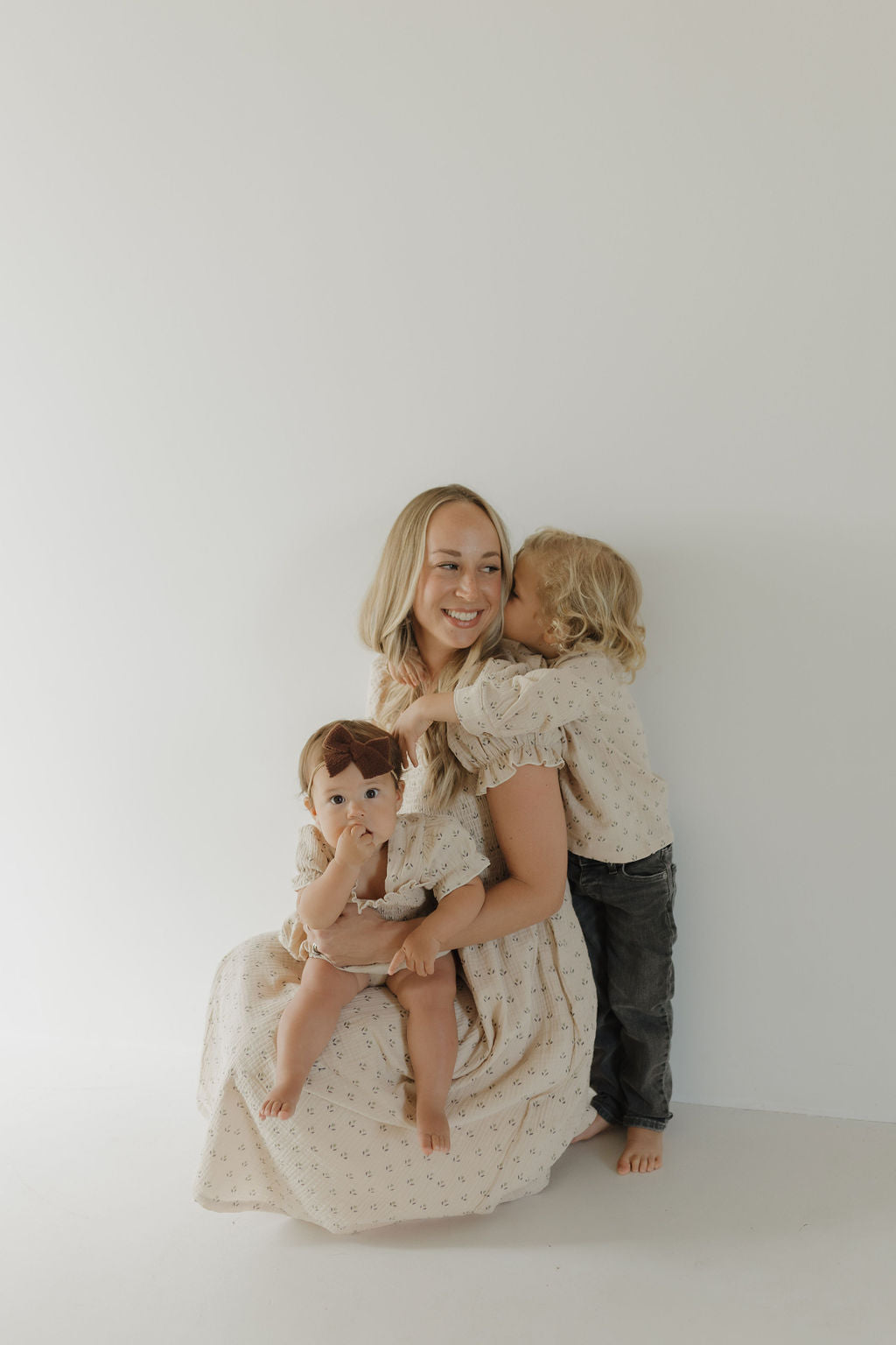 A smiling woman in the forever french baby Women's Short Sleeve Muslin Dress | Flora sits holding a baby, as a blond child hugs her from behind. All three wear matching neutral outfits against a plain white background.